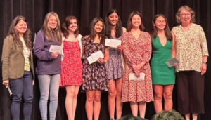 Two teachers and six high school students standing on a stage. All are smiling and students are holding up scholarship awards.