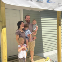 Photograph of a family with mother, father, and 3 children, standing by the front door of a new house. Banner names of donor organizations hangs above their heads.