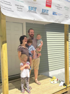 Photograph of a family with mother, father, and 3 children, standing by the front door of a new house. Banner names of donor organizations hangs above their heads.