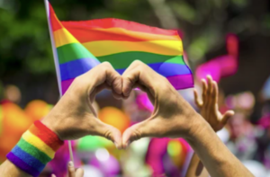 In background, rainbow flag of LGBTQ+ pride; in foreground, two hands forming a heart.