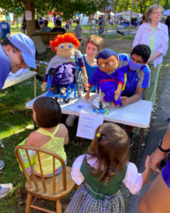 Two children putting on a puppet show for other children. One puppet is in a wheelchair and the other has a white cane such as used by some blind people.