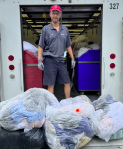 Photograph of man standing in the open back of a truck with a large pile of contributed linens in bags at his feet.