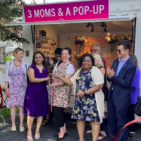 A group of smiling applauding people stand in front of a retail kiosk labeled "3 moms & a pop-up"