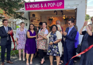 A group of smiling applauding people stand in front of a retail kiosk labeled "3 moms & a pop-up"