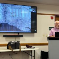 A speaker behind a podium and an aerial terrain map on a video screen.