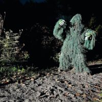 A 'monster' figure in a green ghillie suit against a dark background