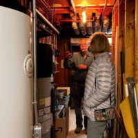 Man and woman in a basement with large tank and heating equipment. Man gesturing.