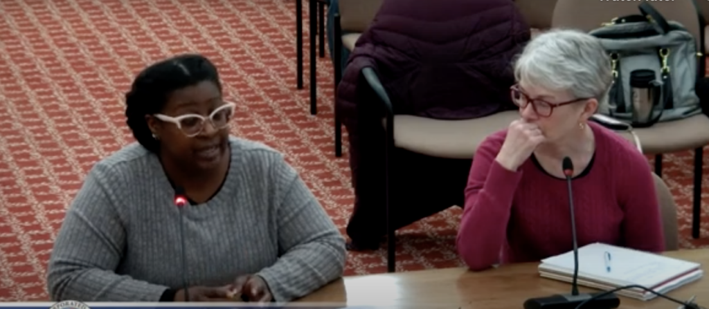 Two women in eyeglasses are seated at a wooden table with microphones. The woman on the left is speaking, as her microphone is lit up. Chairs with coats, and a patterned rug, are in the background.