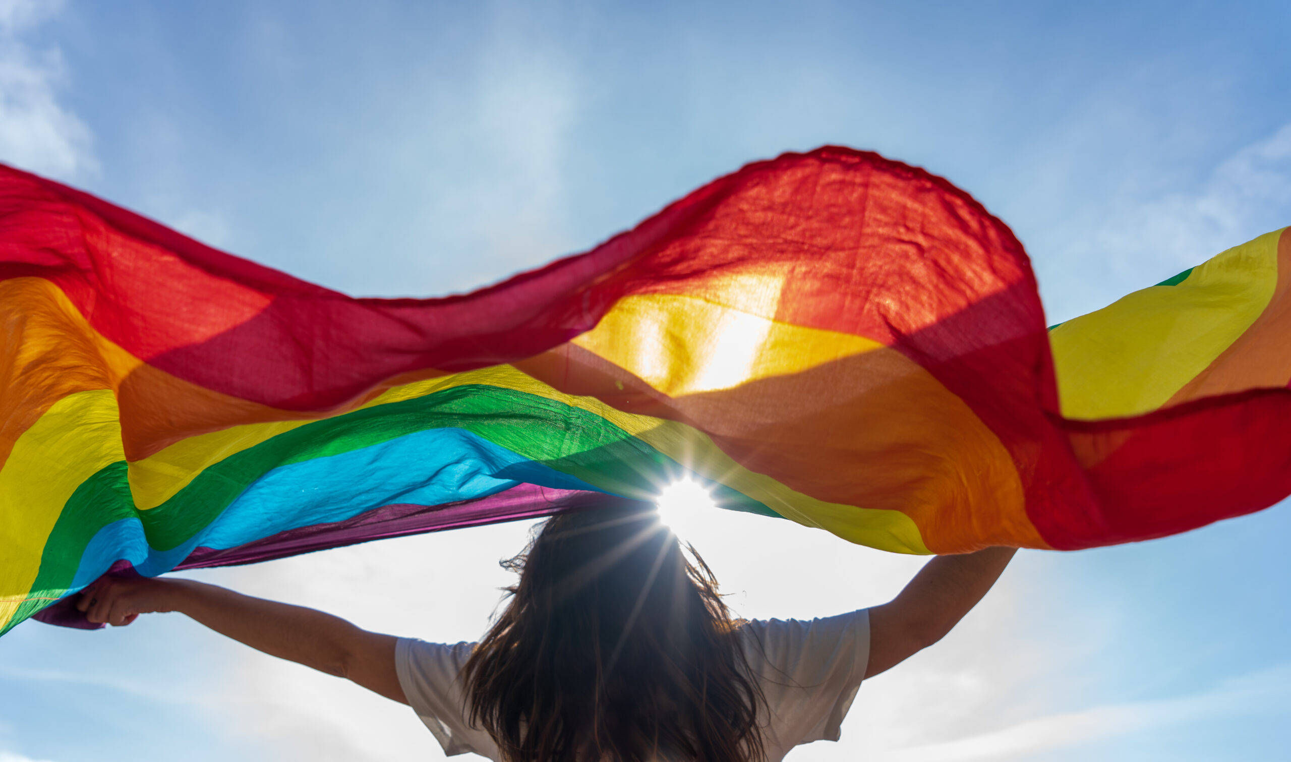 Picture of a young woman waving lgbtq+ flag under the sky