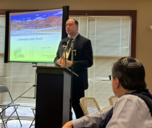 A man stands at a lectern with a video screen in the background