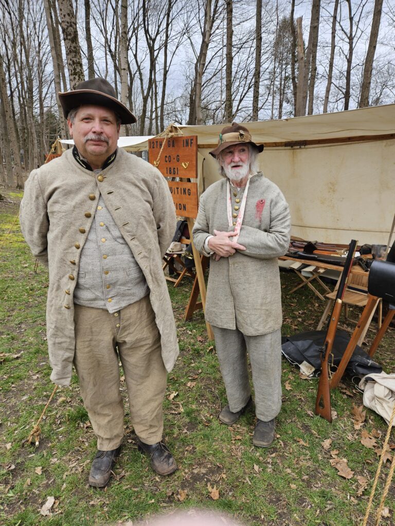 Two men wearing 19th century garb standing in front of a sign that says “12th GA Inf(antry), Co(mpany) G, 1861.”